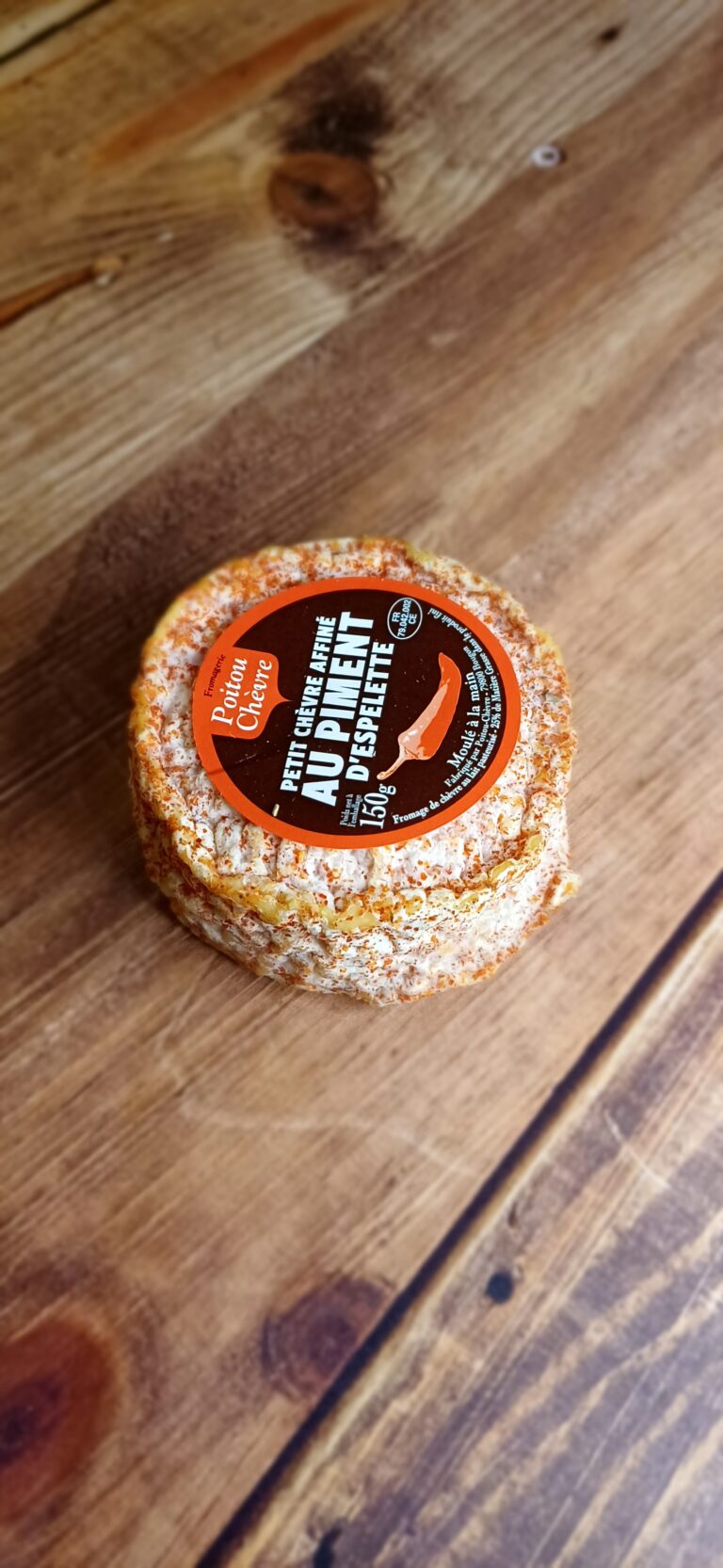 Vue en plongée d'un fromage rond, partiellement enrobé de piment d'espelette. Une étiquette ronde sur le dessus du fromage indique le nom "Chèvre au Piment d'Espelette"